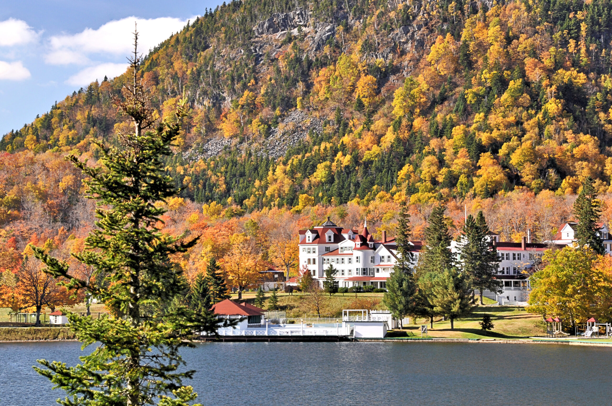 treelined mountainside of colorful fall foliage above lake gloriette and historic balsams grand resort hotel in remote dixville notch, new hampshire.